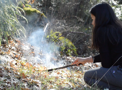 Person crouching in a forested area, using a stick to tend a small, smoky ground fire among dry leaves and grass.
