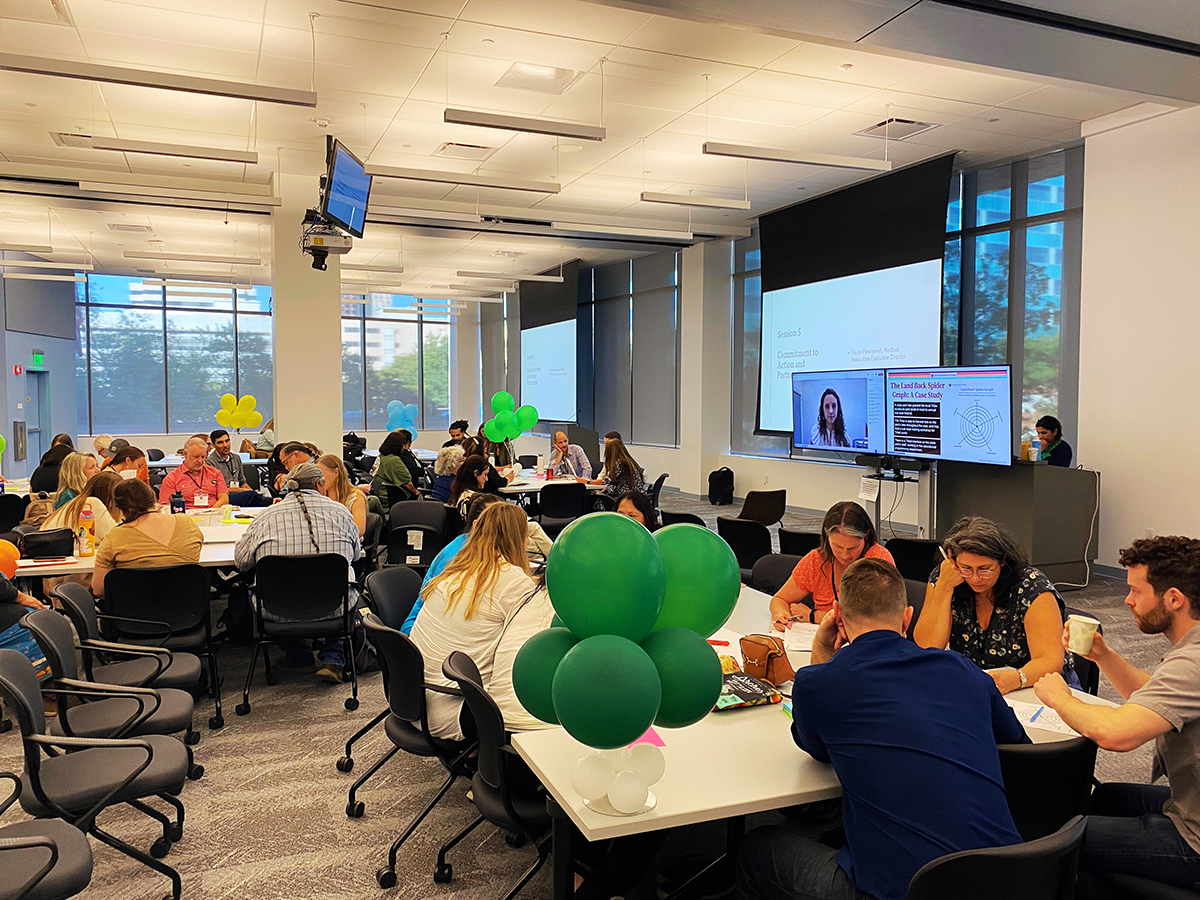 Large conference room with multiple groups of people seated at tables in discussion, green balloons in the foreground and a presentation with a remote speaker displayed on screens at the front of the room.