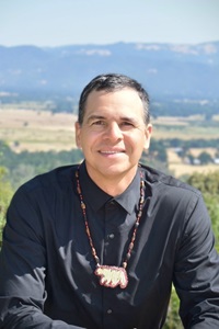 Headshot of Professor William Bauer Jr., Round Valley Indian Tribes citizen and UC Riverside history professor, wearing a black shirt and beaded necklace outdoors with mountains in the background.