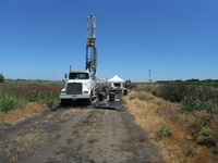 Photo of truck-mounted drill rig with support trucks and equipment used for soil exploration. A temporary tent structure by the drill rig is used to process soil samples and provide shade for the working crew.